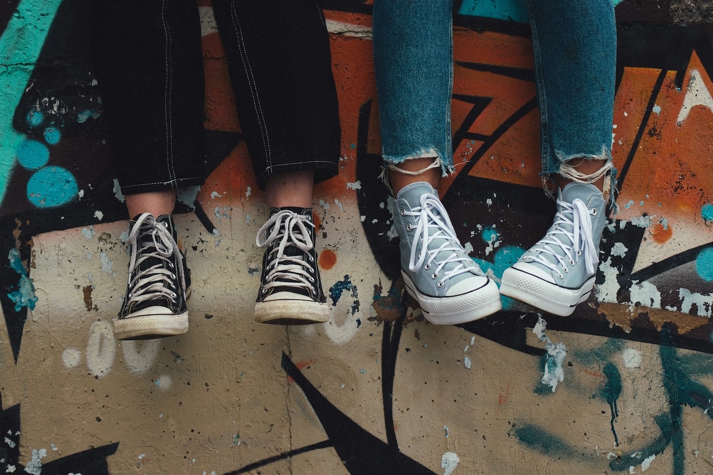 Two teens stand side by side on a graffiti-painted floor, shown from the knees down in jeans and canvas sneakers.