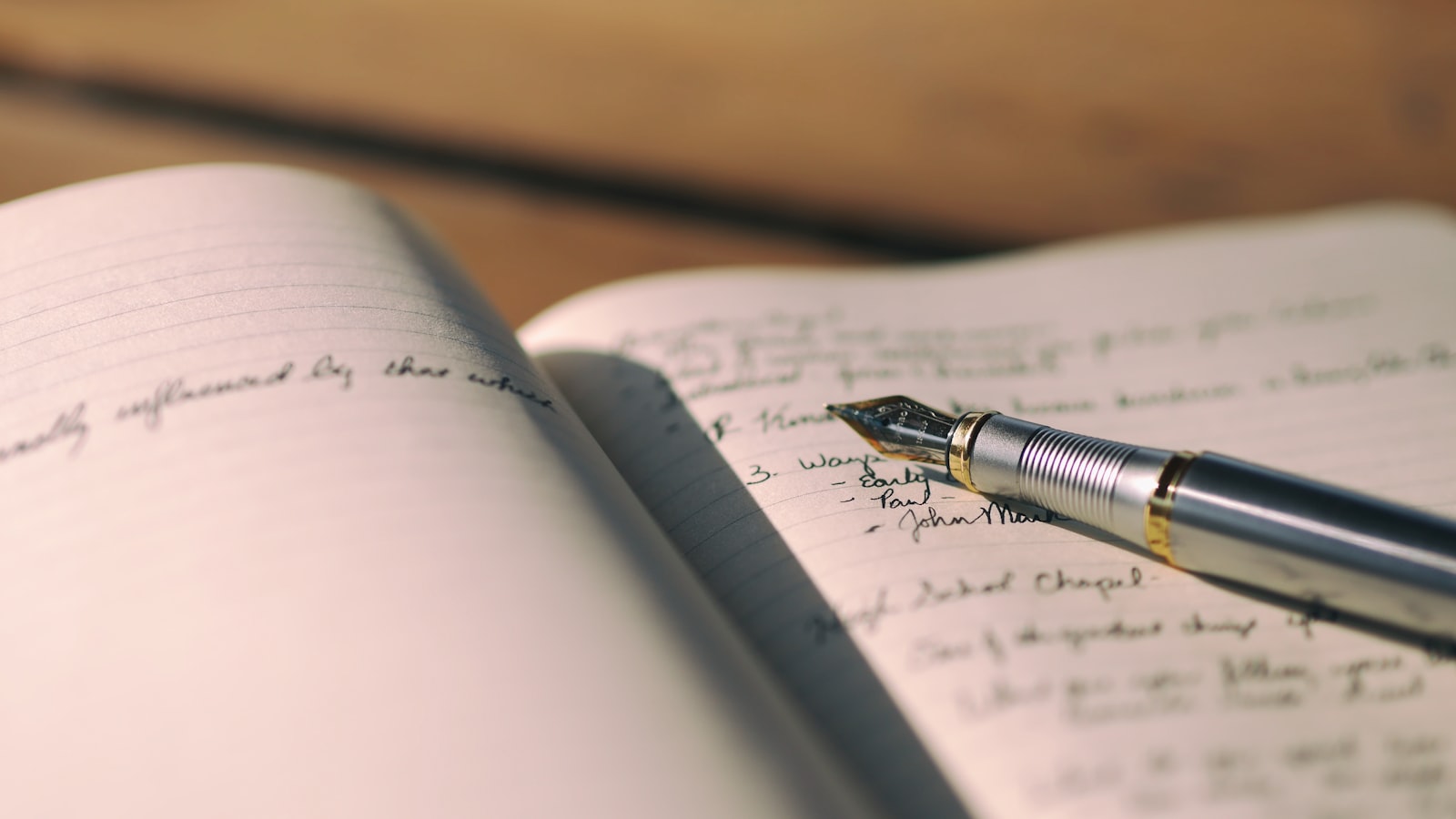 A fountain pen rests on the open ruled page of a paper notebook in soft natural light.