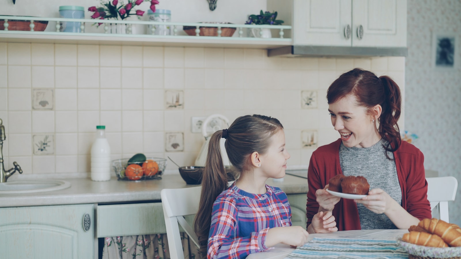A mother shares a basket of fresh muffins with her daughter at a warm-lit kitchen table.