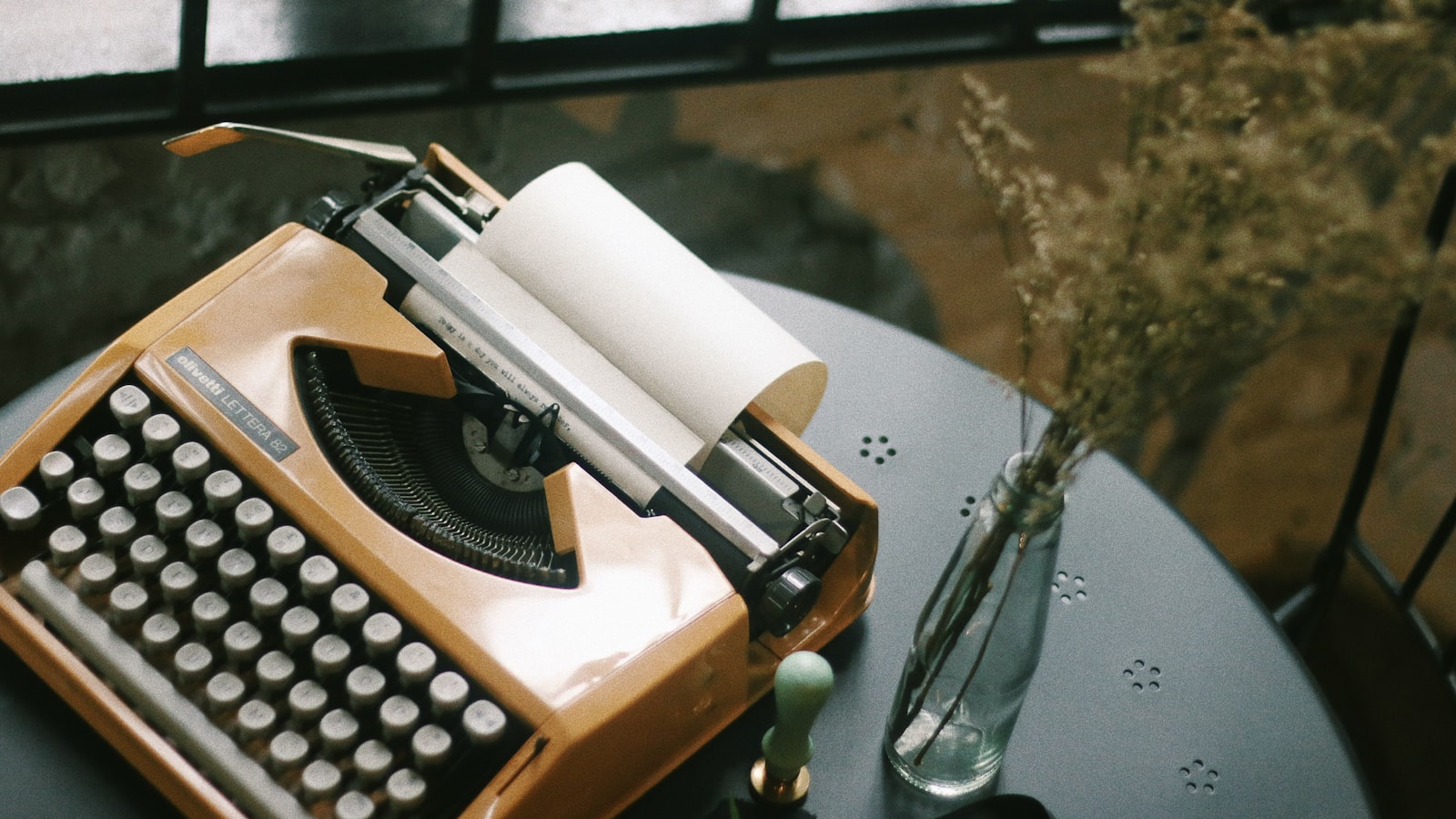 A vintage typewriter and a sprig of dried flowers on a table by a window in soft natural light.